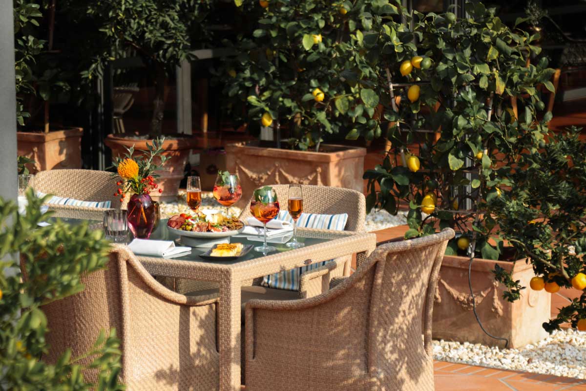 A patio dining table set for four with drinks and colorful dishes, surrounded by potted lemon trees and greenery in an outdoor garden setting.