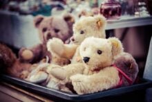 Several plush teddy bears, mostly beige and brown, sit together in a black container at what appears to be a market or sale. The background shows blurred objects and other items on display.