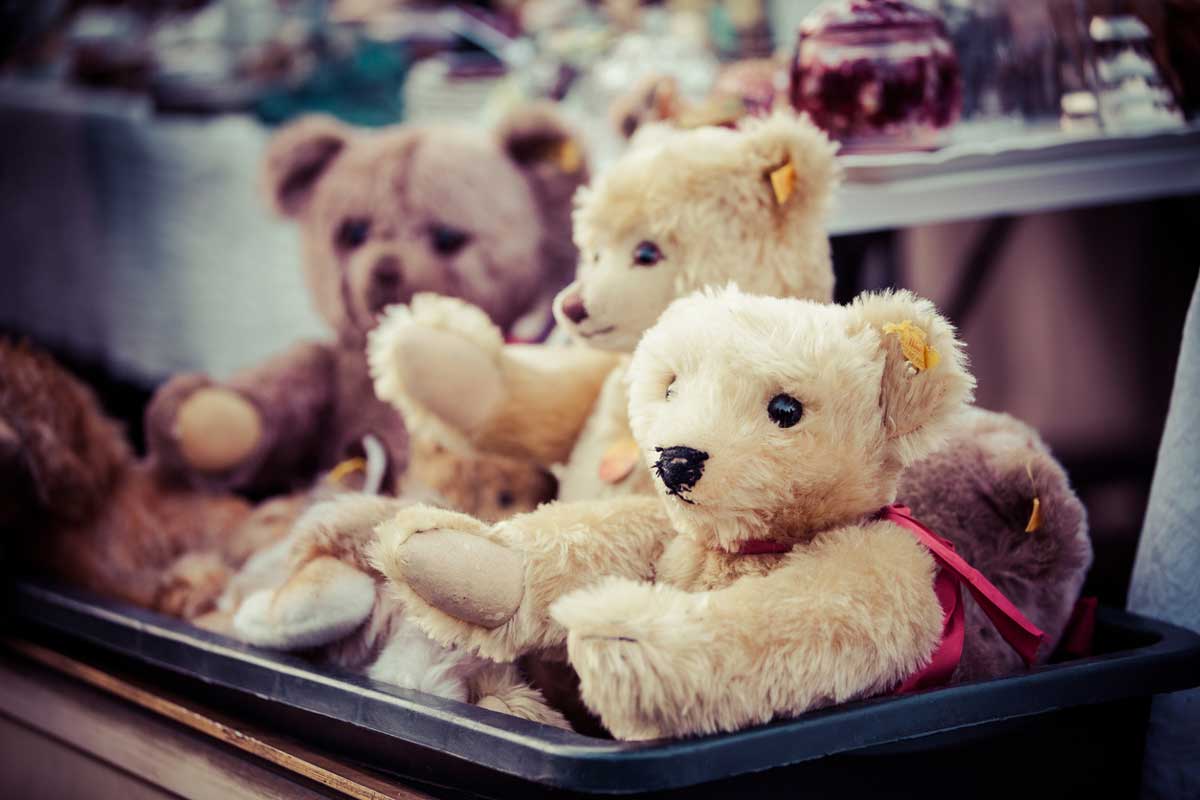 Several plush teddy bears, mostly beige and brown, sit together in a black container at what appears to be a market or sale. The background shows blurred objects and other items on display.