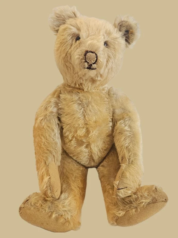 A vintage, light brown teddy bear with worn fur, dark stitched nose and mouth, and beige fabric on its paws and feet, sitting upright against a plain beige background.
