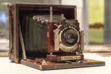 A vintage Korona II.B folding camera is displayed on a table. The camera features a bellows design and a wooden frame, with metal components visible. It is positioned upright, showcasing its classic and intricate mechanical details.