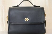 A black leather handbag with a top handle, gold turn-lock clasp, and structured shape, set against a light background.