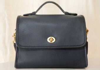 A black leather handbag with a top handle, gold turn-lock clasp, and structured shape, set against a light background.