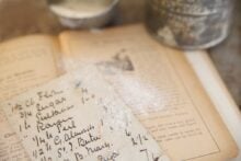 A handwritten recipe card dusted with flour rests on an open cookbook, with a metal sifter and a bowl in the background, evoking a vintage, homey baking scene.