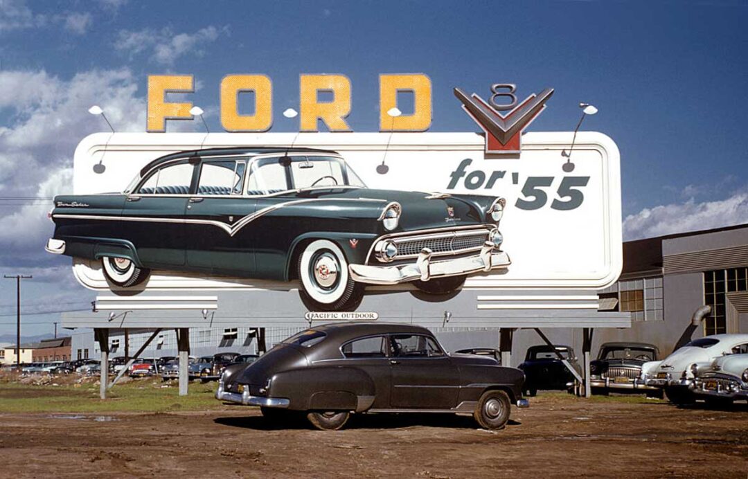 A large roadside billboard advertises the 1955 Ford car, showing a green and white model. Below the sign, an older black car is parked on a dirt lot with several other parked cars and an industrial building in the background.