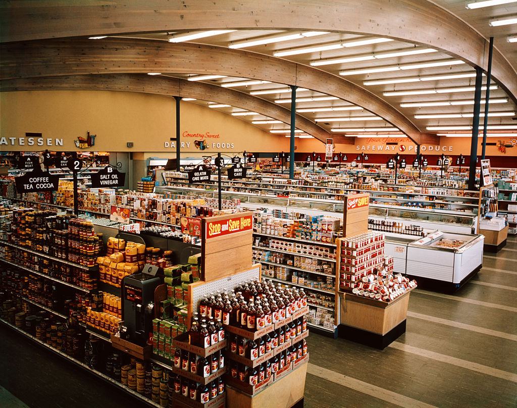 Wide view of a retro supermarket with curved wooden ceiling and bright lighting. Aisles are stocked with various products like jars and bottles. Signs indicate sections for deli, dairy, and produce. Customers and carts are visible throughout.