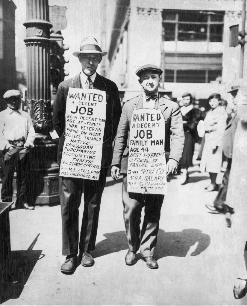 Two men stand on a busy street wearing sandwich boards with handwritten messages seeking jobs. The man on the left is taller and wears a hat. The man on the right has a shorter build and is dressed in a suit and cap. People walk by in the background.