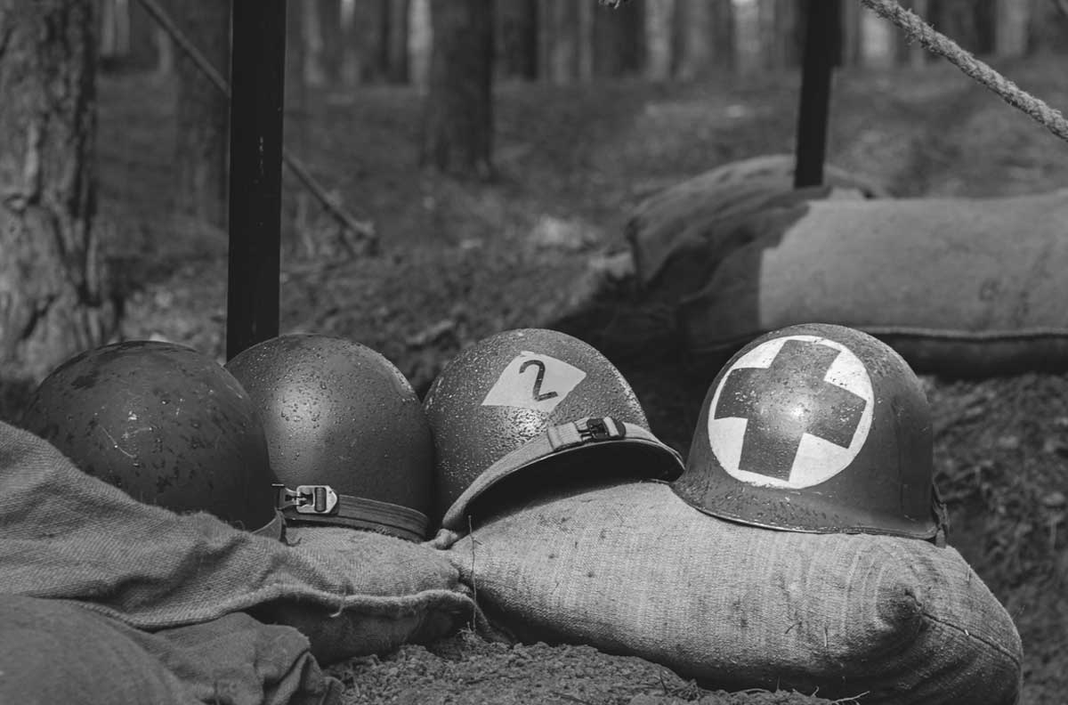 Four military helmets rest on sandbags in the woods; one helmet displays a medical cross. The scene is in black and white, evoking a historical wartime setting.