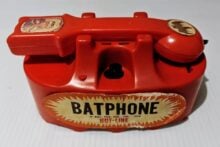 A vintage red toy phone labeled "Batphone" with a bat symbol and the words "Hot-Line." The phone features retro design elements and is from 1966.