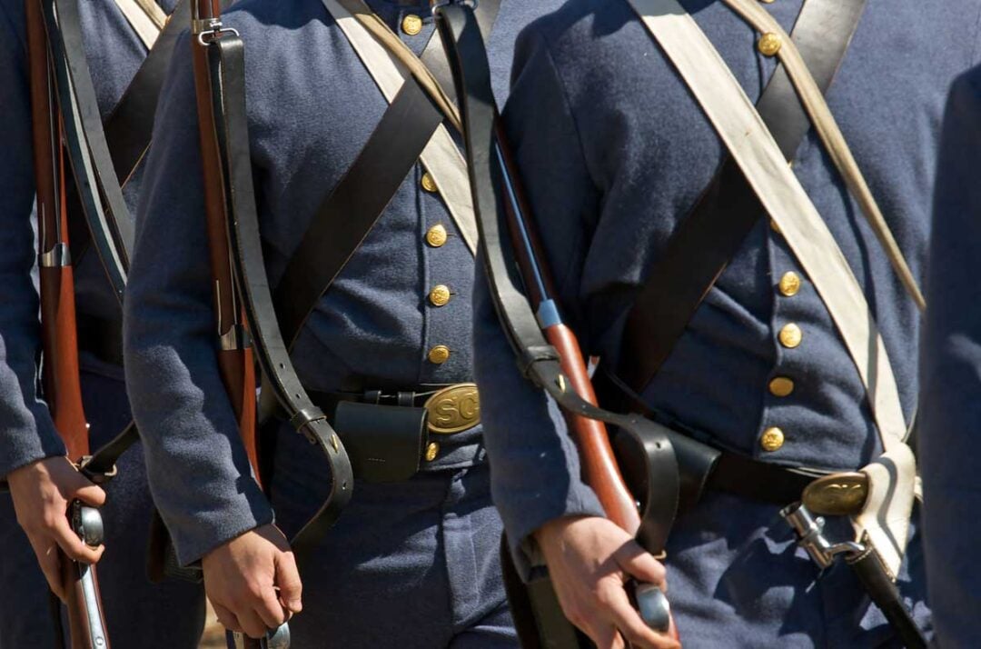 Several people in blue Civil War-era military uniforms with brass buttons and white straps march with rifles on their shoulders, visible from the chest down.