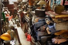 A cluttered antique shop filled with stacks of old fabrics, pottery, lamps, books, and various vintage items, all piled closely together along narrow aisles.