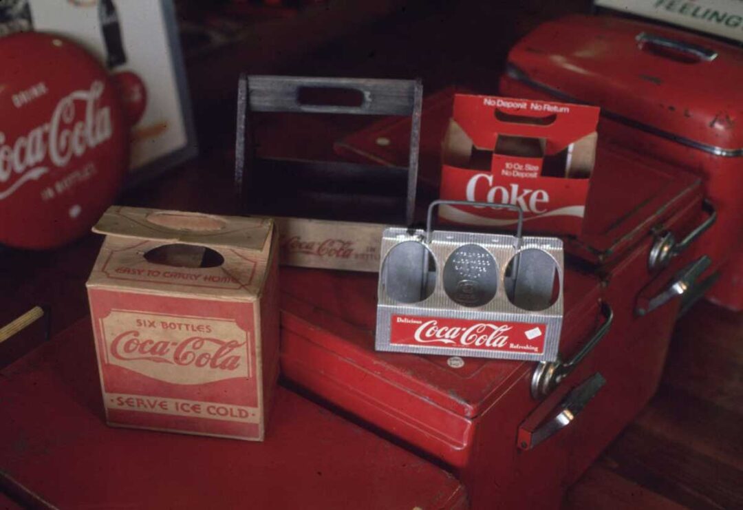 Vintage Coca-Cola and Coke bottle carriers sit on red suitcases, including cardboard and metal designs for holding six bottles, with retro branding and slogans visible.