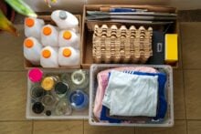 Three containers are organized for recycling: one holds empty plastic bottles, another contains egg cartons, papers, and small boxes, and the third has clean, folded clothes and glass jars on a tiled floor.