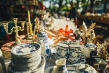 A table at an outdoor flea market displays vintage blue and white china, cups, saucers, a teapot, brass candlesticks, and various decorative items in sunlight.