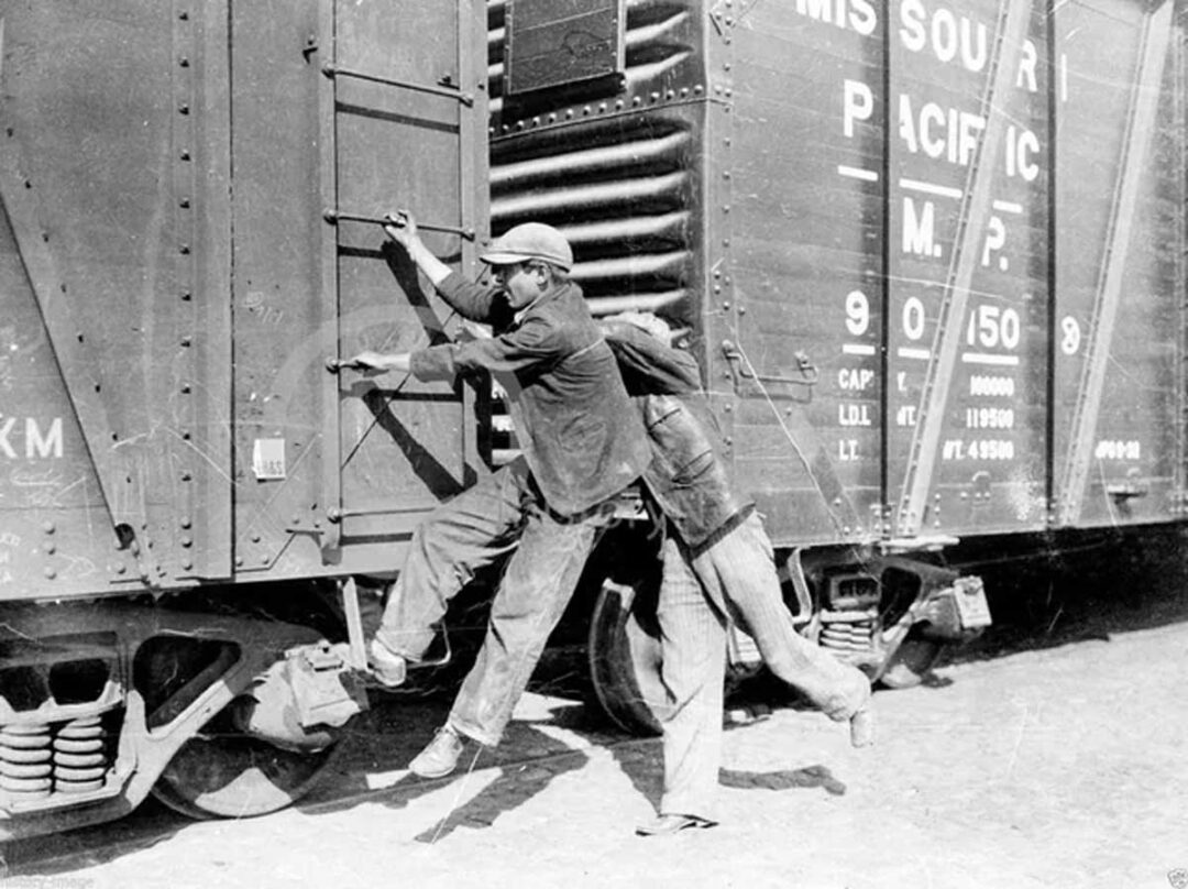 Two men are jumping onto a moving freight train. One man grabs a ladder while the other pushes from behind. The train has visible text reading "Missouri Pacific" and appears weathered and industrial.