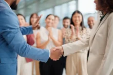 Two people shake hands in focus while a group of diverse individuals in the background applaud and smile, suggesting a successful agreement or celebration. They are in a bright, modern indoor setting with large windows.