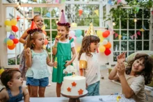 A group of children wearing party hats celebrate at an outdoor birthday party. They're gathered around a decorated cake with lit candles. Colorful balloons and bunting adorn the background. One adult laughs and claps nearby.