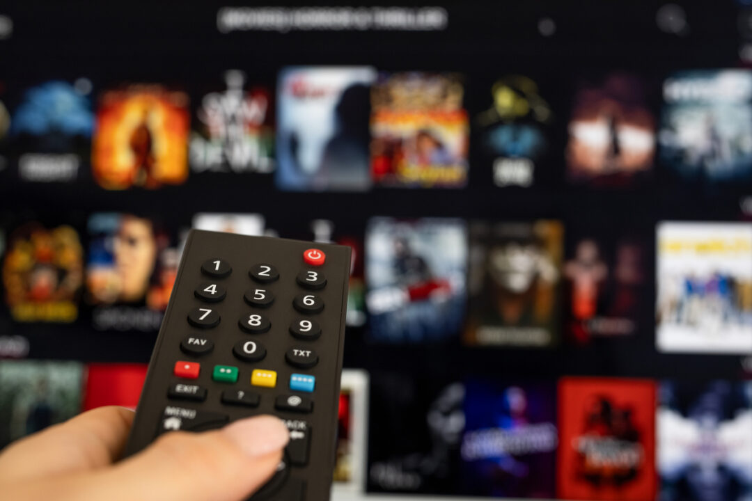 A hand holds a remote control in front of a large screen displaying a streaming service interface. Various movie and TV show thumbnails are visible, blurred in the background.