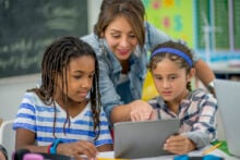 A teacher smiles and helps two students, one wearing a striped shirt and another with a headband, as they look at a tablet together in a classroom. There's a chalkboard and colorful posters in the background.
