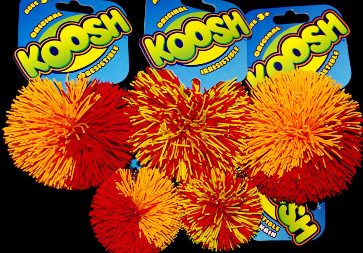 Five red and yellow Koosh balls are displayed in front of blue packaging that features the yellow “Koosh” logo. The colorful balls are made of soft rubber strands and arranged against a black background.