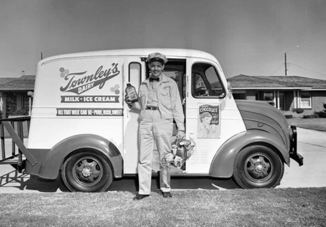 A smiling milkman in uniform stands beside a Townley’s Dairy delivery truck, holding a milk bottle and a basket, in a suburban neighborhood. The truck advertises milk and ice cream.
