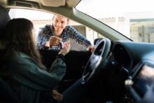 A man standing outside a car smiles and hands a car key to a woman sitting in the driver&rsquo;s seat, who is reaching out to take it.