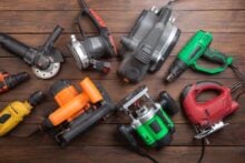 A variety of electric power tools, including a jigsaw, drill, heat gun, nail gun, grinder, and router, arranged on a wooden surface. The tools are in different colors and positions.