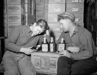 Two men sit in a warehouse with stacks of boxes. One man leans against a box, examining a whiskey bottle. Several other whiskey bottles are displayed on the box. Both are wearing work clothes and caps, with more boxes seen in the background.