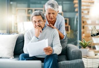 An older man sits on a couch, reading and looking concerned at papers in his hand, while an older woman stands behind him with a supportive hand on his shoulder. A laptop and plants are on the table nearby.