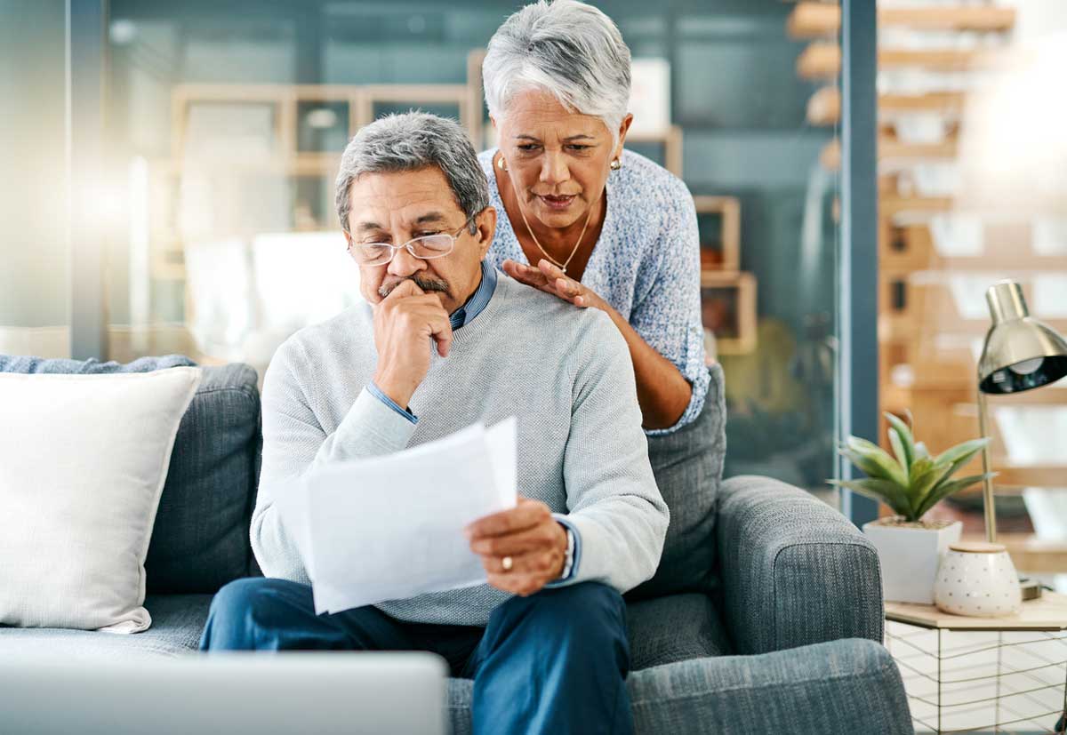 An older man sits on a couch, reading and looking concerned at papers in his hand, while an older woman stands behind him with a supportive hand on his shoulder. A laptop and plants are on the table nearby.