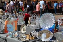 A table at an outdoor market displays vintage silverware, glass decanters, crystal dishes, china, and a colorful portrait. Shoppers and vendors are seen blurred in the background.