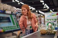 A woman with long red hair stands at a grocery store checkout, unloading a pineapple and milk onto the conveyor belt, surrounded by brightly lit shelves.