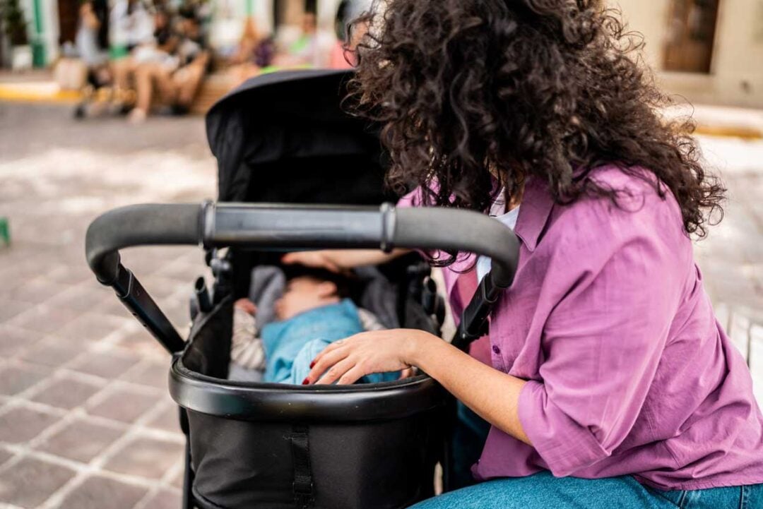 A woman with curly hair and a purple shirt sits outdoors, gently touching a baby lying in a black stroller on a cobblestone street.