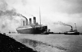 A large ocean liner with four smokestacks, emitting smoke, sails near smaller boats on a wide river under an overcast sky. People stand on the riverbank in the foreground.