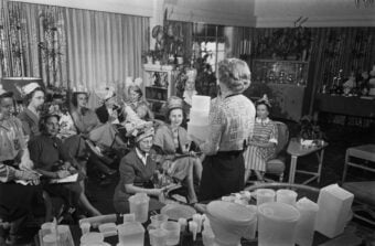 A woman stands in front of a group, holding plastic containers, demonstrating their use to an audience of women seated in a living room decorated with plants and furniture.