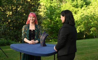 A person with pink hair and a patterned shawl stands behind a blue table with a jewelry display. Another person in a dark suit listens attentively. They are outdoors, with lush greenery in the background.