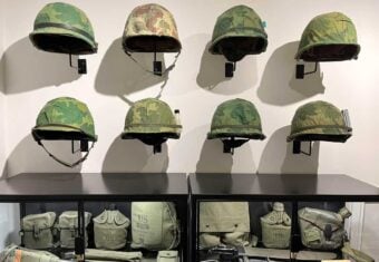 Nine military helmets with camouflage covers are mounted on a wall above two glass display cases containing various green military gear, including canteens, pouches, and bags, all labeled "US.