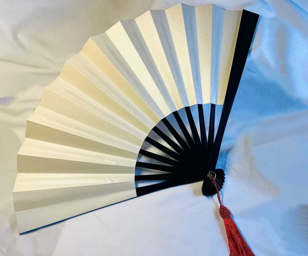 A traditional Japanese folding fan with white paper and black ribs, decorated with a red tassel, displayed on a white fabric background.