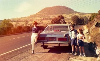 A woman and two children stand by a parked car on the side of a rural road, with a sunlit hill and trees in the background. The atmosphere is relaxed and nostalgic.