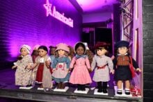 A group of American Girl dolls is displayed on a stage. The dolls are dressed in various historical outfits, and a pink neon "American Girl" sign is illuminated in the background.
