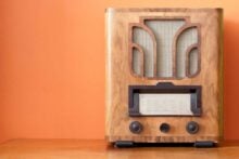 A vintage wooden radio with a fabric speaker grille and two dials sits on a wooden surface against an orange wall.