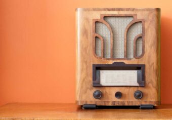 A vintage wooden radio with a fabric speaker grille and two dials sits on a wooden surface against an orange wall.