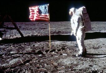 Astronaut in a white spacesuit stands on the moon's surface beside an American flag. The lunar module is partially visible in the background, and footprints mark the gray, dusty ground under a black sky.
