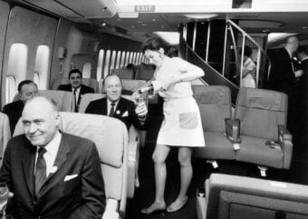 A flight attendant in a classic uniform pours a drink for a seated male passenger in the first-class cabin of a vintage airplane, with other well-dressed passengers seated nearby.