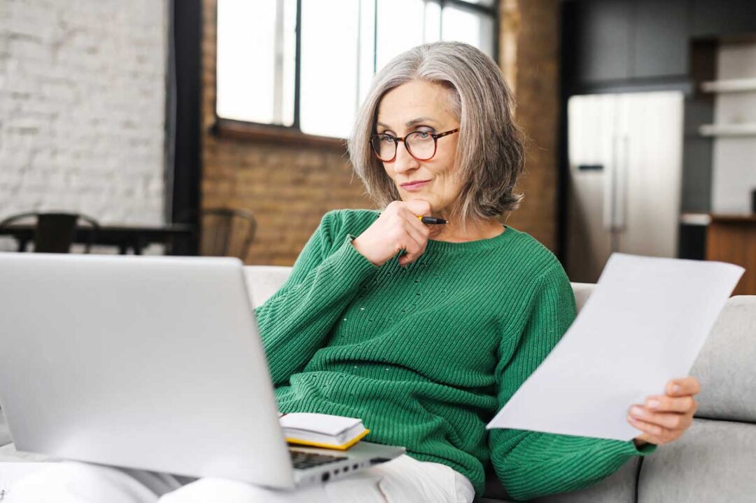 An older woman with gray hair and glasses sits on a couch, holding a paper in one hand and a pen near her chin, looking thoughtfully at a laptop. She is wearing a green sweater and appears to be working from home.
