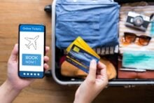 A person holds a smartphone displaying an online flight ticket booking app and two credit cards in front of an open suitcase packed with clothes, a camera, sunglasses, and travel essentials.