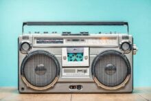A vintage silver boombox with large speakers, cassette deck, and radio tuner sits on a wooden surface against a blue background.