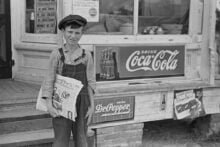 A young boy wearing a cap and overalls stands outside a store holding newspapers. Behind him are vintage Coca-Cola, Dr Pepper, and spark plugs signs displayed on the building.