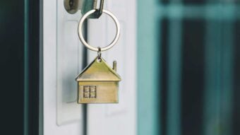 A close-up of a key with a house-shaped keychain inserted into a door lock, symbolizing home ownership or real estate. The background is softly blurred.