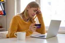A woman in a yellow sweater sits at a table holding a credit card and looking worriedly at a laptop. A white mug is beside her. She appears to be having difficulty with an online transaction.